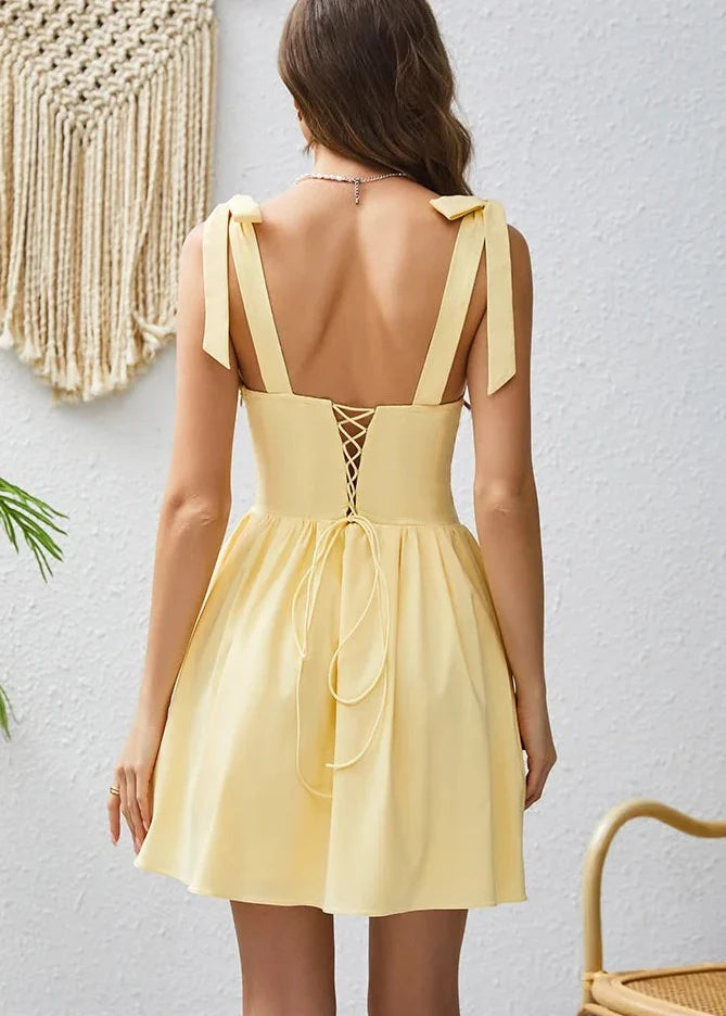 Woman wearing a yellow dress with cut-out back design, standing against a white wall.