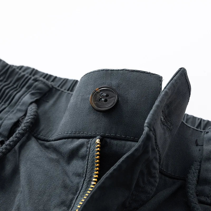 Close-up of dark grey shorts showing a tortoiseshell button, brass zipper, and elastic drawstring waistband against a white background.