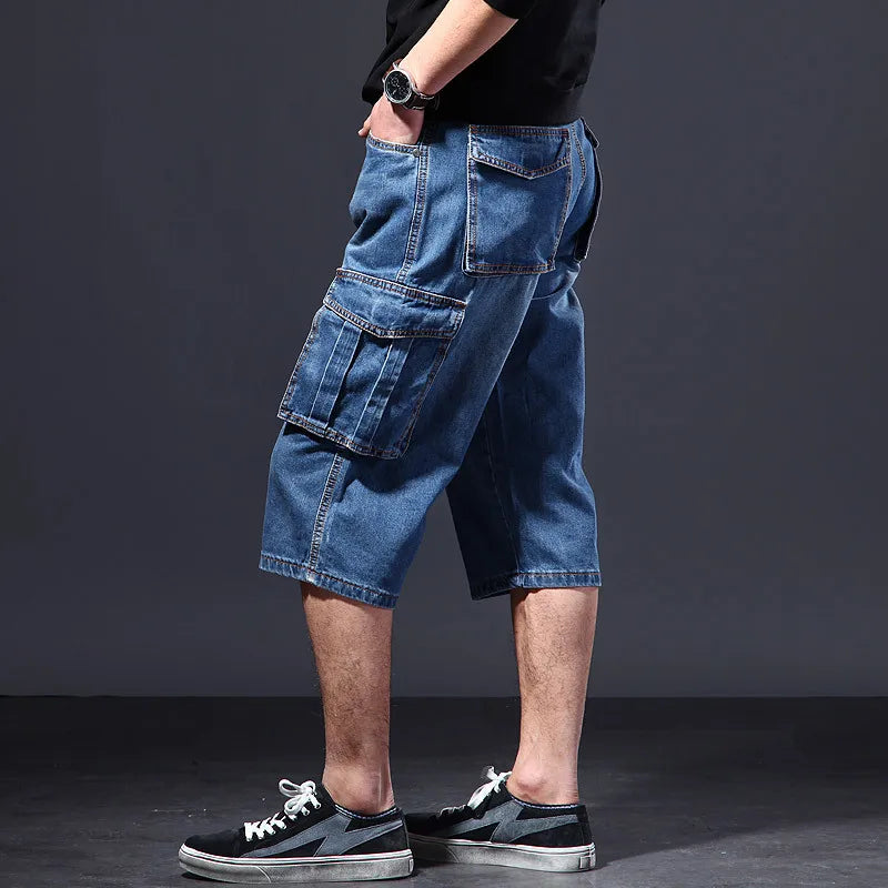 Man wearing blue denim cargo shorts with large pleats on the side cargo pockets, standing against a dark background.
