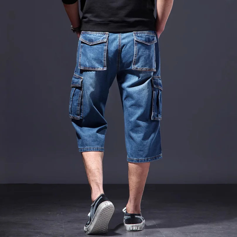Man wearing loose-fitting blue denim cargo shorts with large flap pockets on the sides and back, viewed from behind.