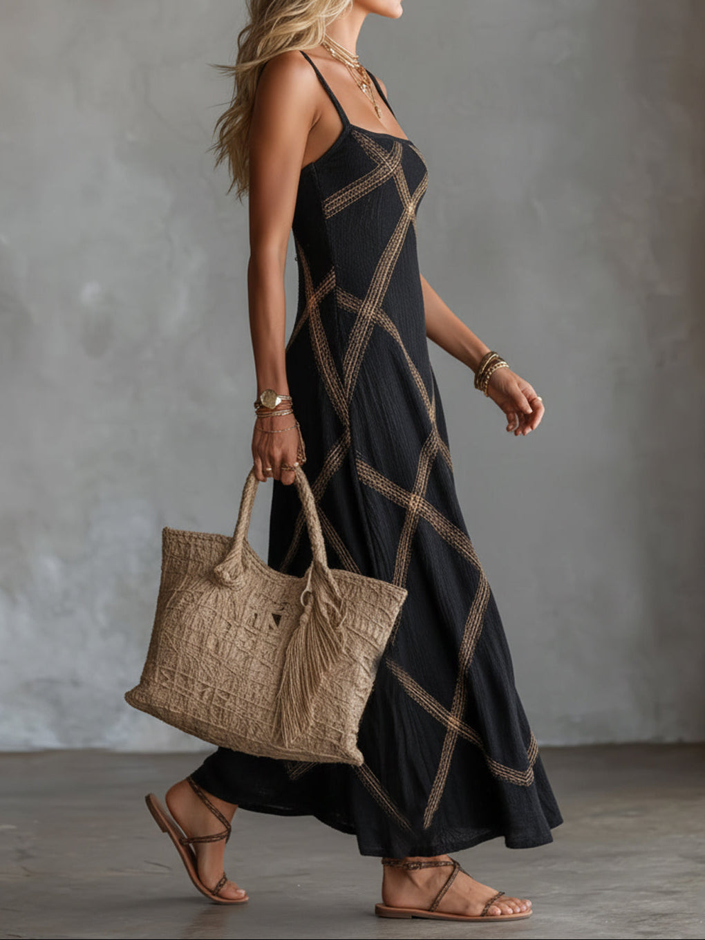 Woman walking in a black maxi dress with gold crisscross pattern, carrying a large natural woven bag with tassels.