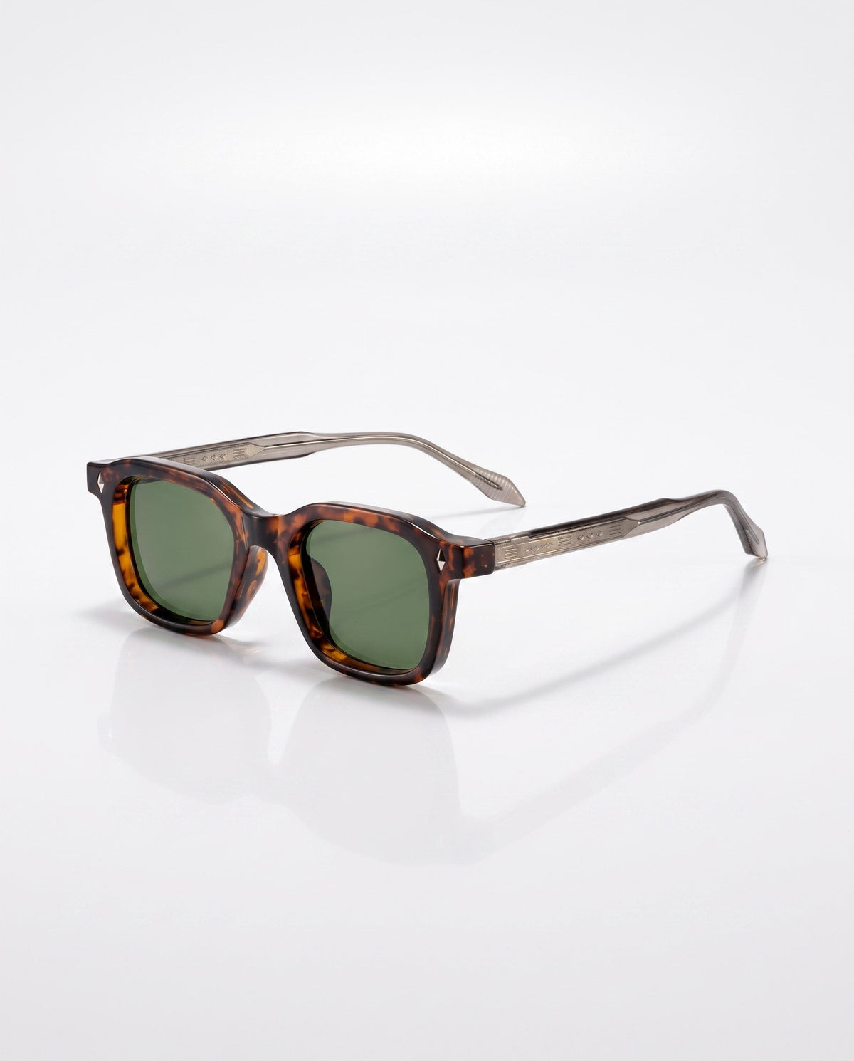 Square tortoiseshell sunglasses with green lenses and patterned metallic temples are angled on a white surface.