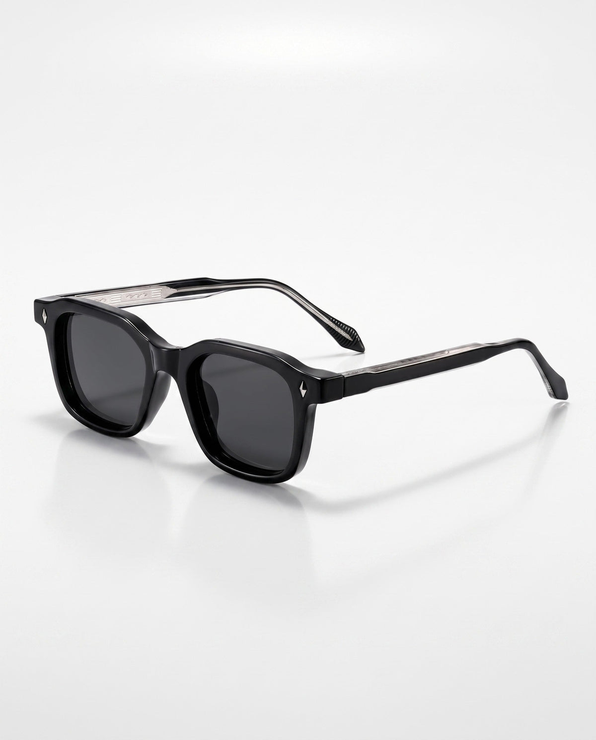 Black square-framed sunglasses with dark lenses and subtle silver diamond accents are angled on a white surface.