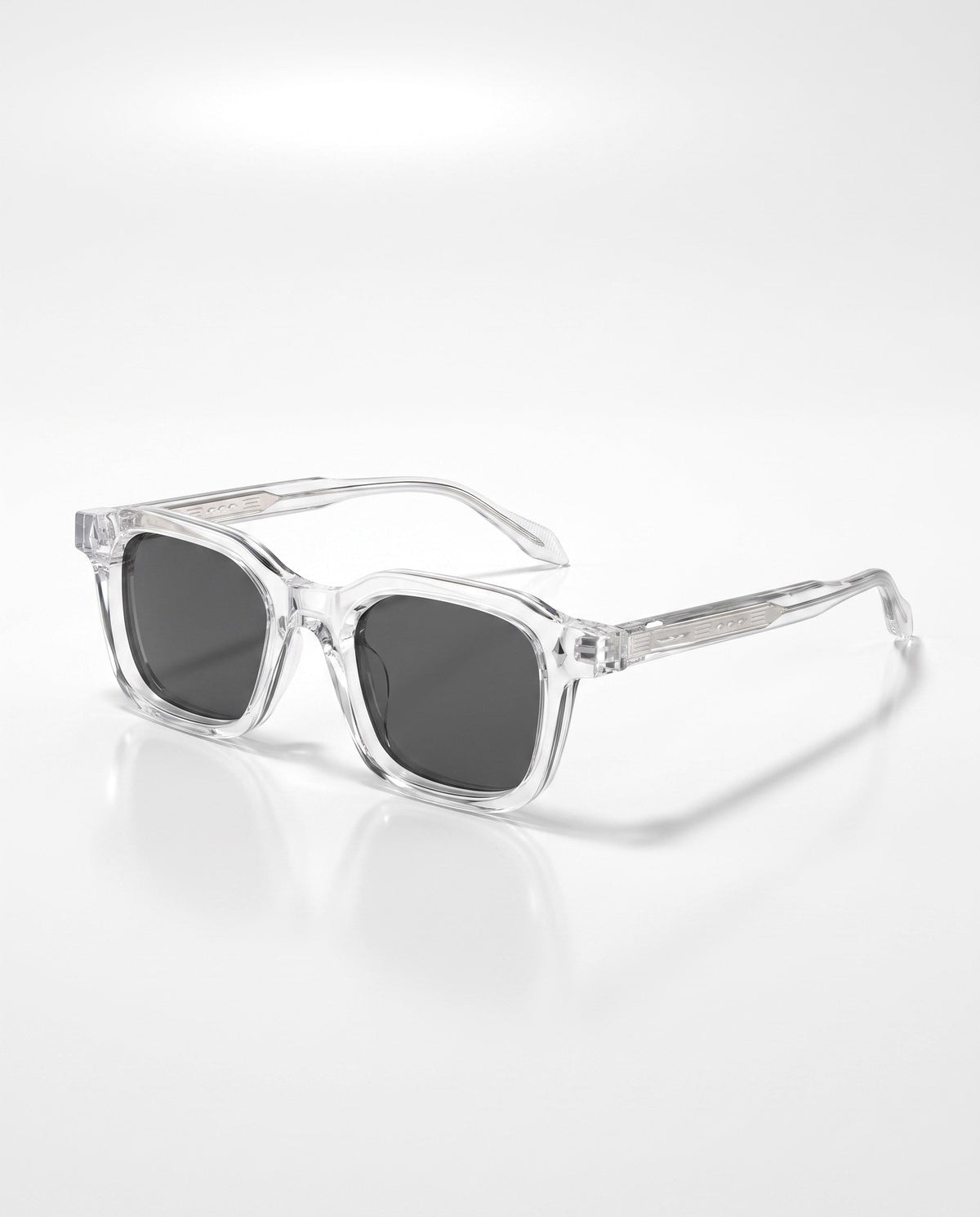 Clear, transparent square-framed sunglasses with dark lenses lie angled on a white surface, casting a soft shadow.