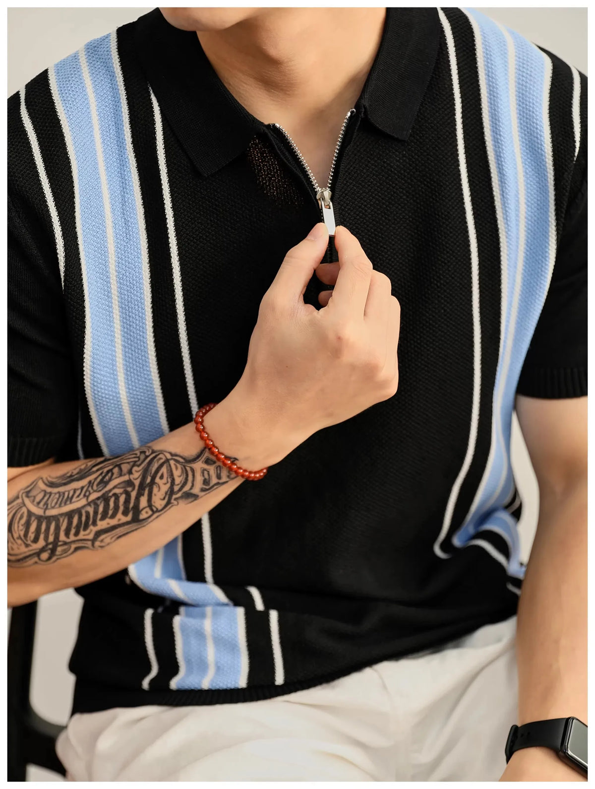 Man wearing a black short-sleeved knit shirt with light blue and white vertical stripes, adjusting the silver zipper.