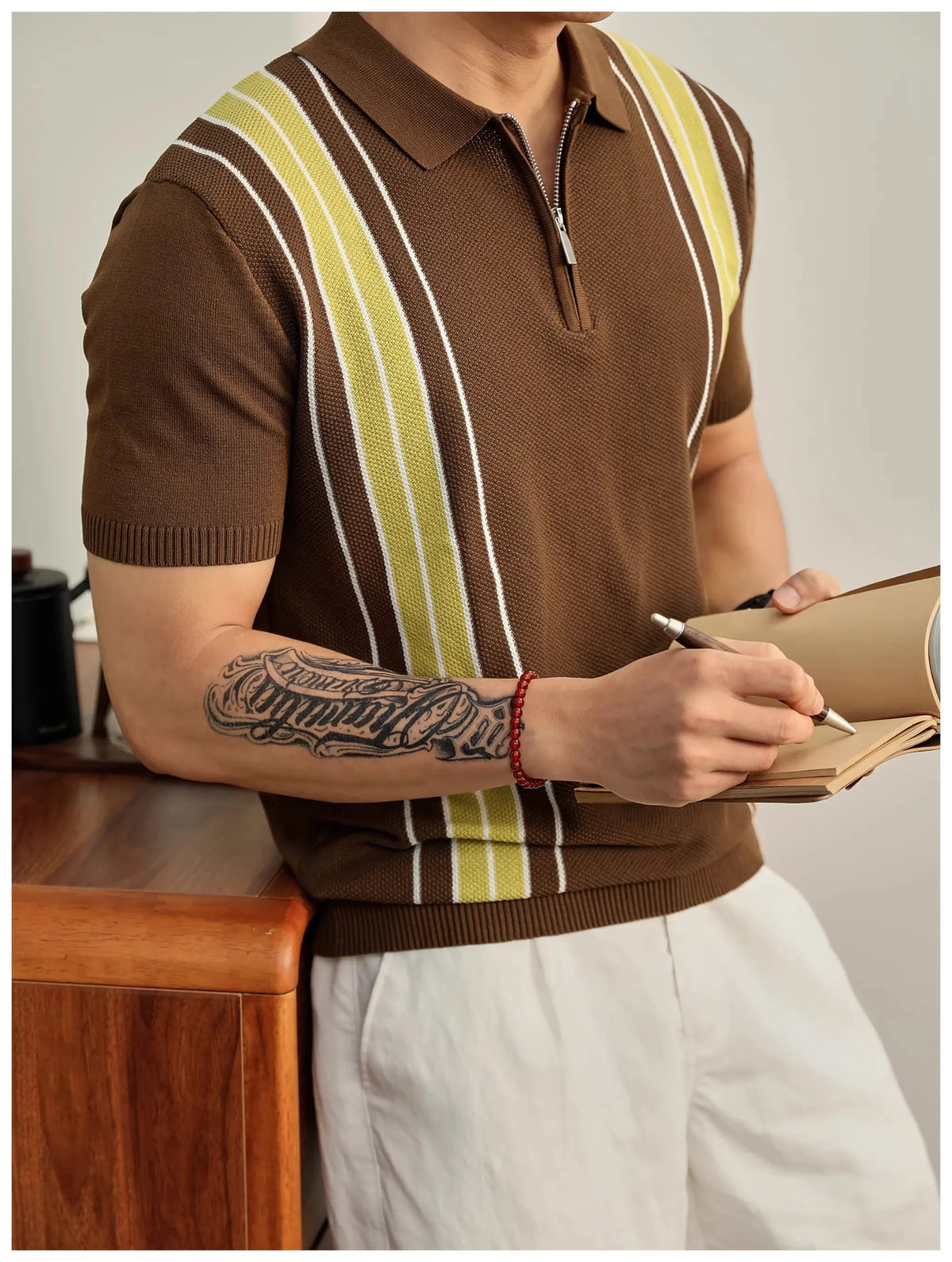 Man wearing a brown knit polo shirt with yellow and white vertical stripes, holding a notebook and pen, against a light background.