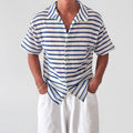 Man wearing a white and blue striped short-sleeve knit shirt with a collar, hands in white pants pockets.