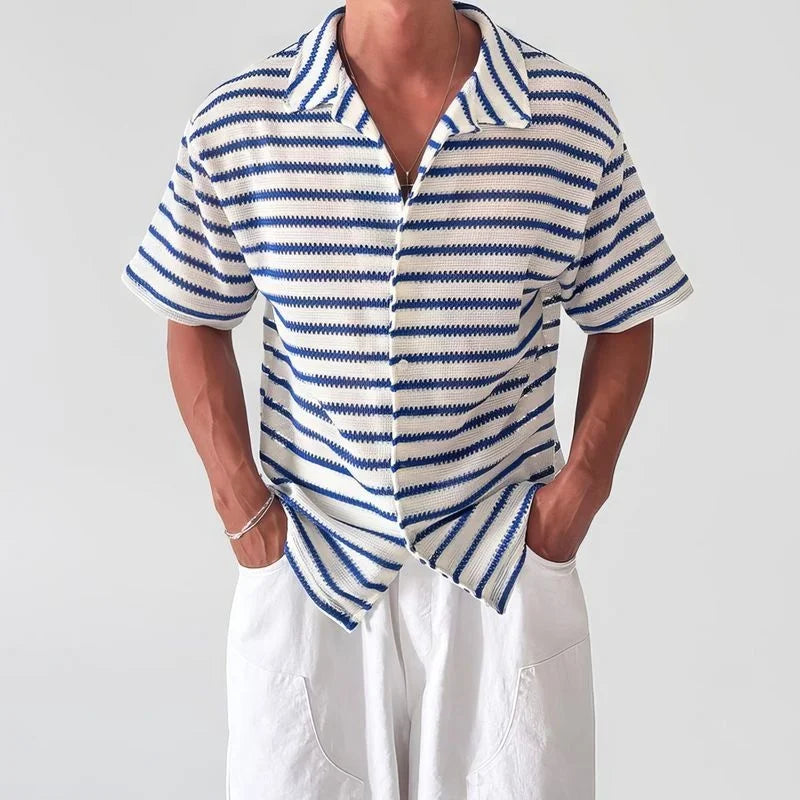 Man wearing a white and blue striped short-sleeve knit shirt with a collar, hands in white pants pockets.
