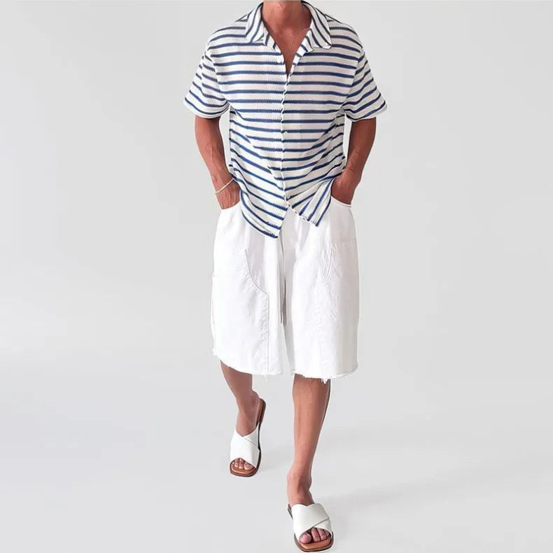 Man wearing a short-sleeve collared shirt with horizontal navy and white stripes, paired with white shorts and sandals.