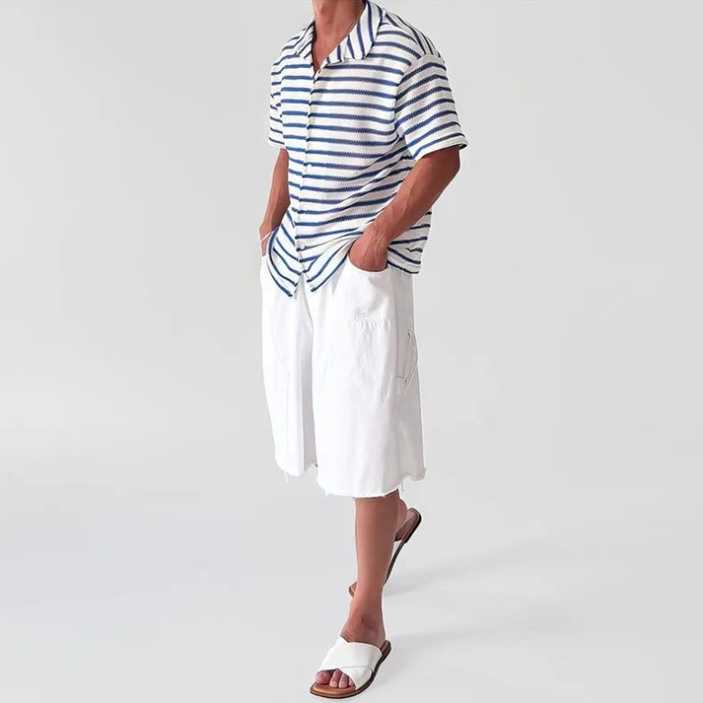 Man wearing a loose-fitting, short-sleeve knit shirt with blue and white stripes, paired with white shorts and sandals.