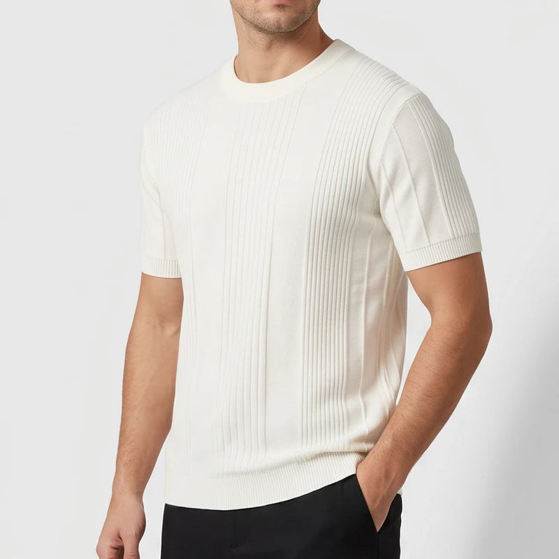 Man wearing a cream ribbed short-sleeve knit shirt with a crewneck, standing with one hand in a black pant pocket.