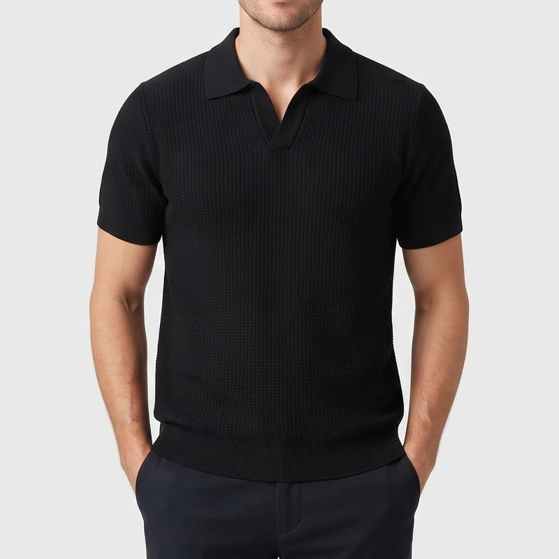 Man wearing a black short sleeve knit polo shirt with a textured pattern and an open collar, hands in dark pants pockets.