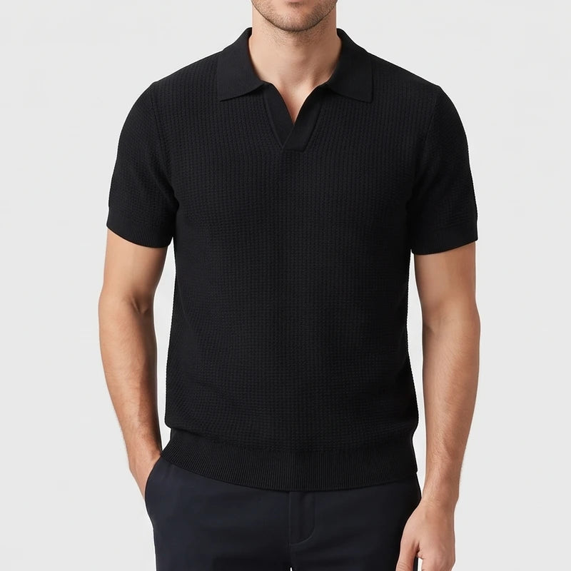 Man wearing a black short sleeve knit polo with a textured waffle pattern, an open collar, and ribbed hem, standing against a light background.