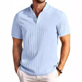 Man wearing a light blue short-sleeve textured shirt with a band collar and two white buttons, hands in white pants pockets.