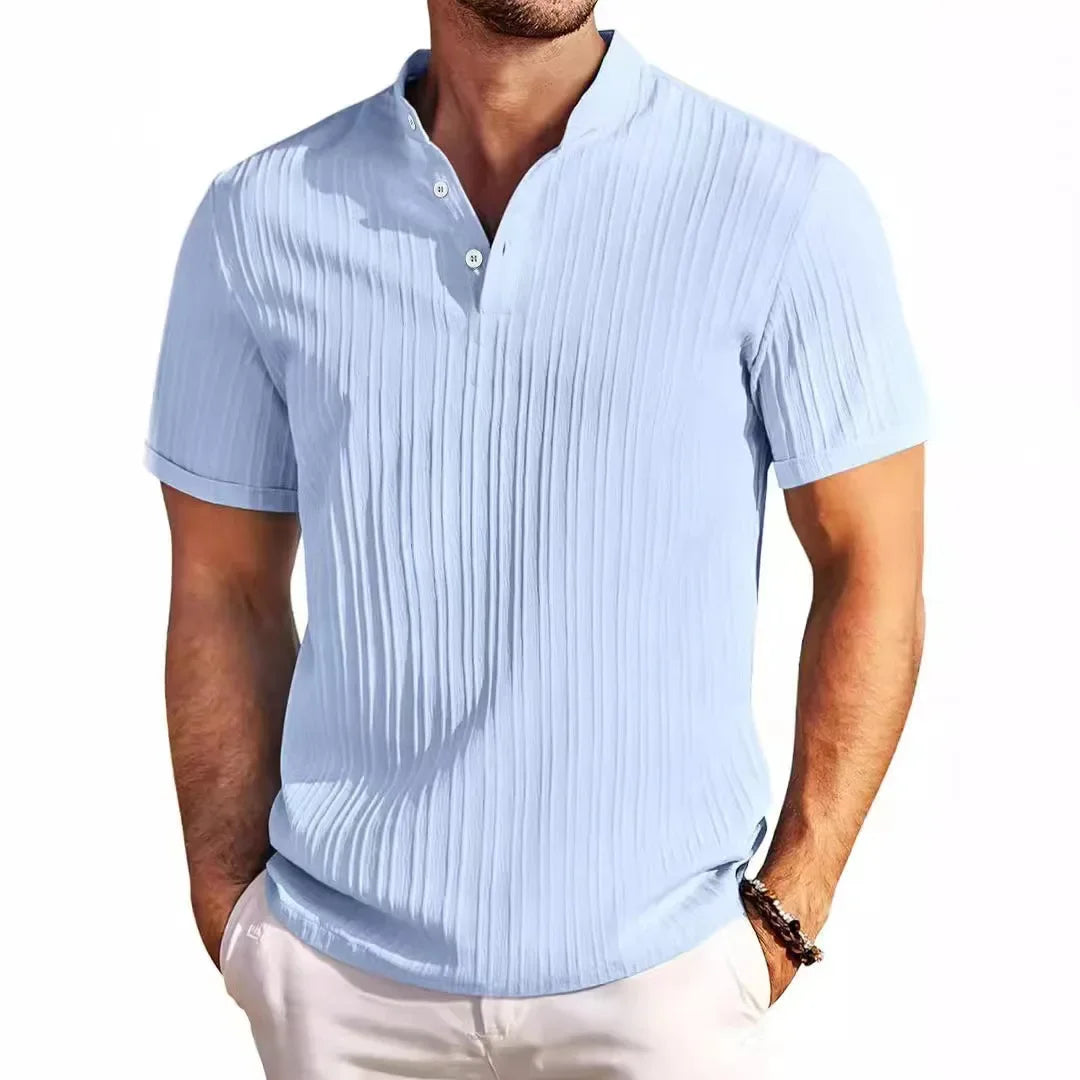 Man wearing a light blue short-sleeve textured shirt with a band collar and two white buttons, hands in white pants pockets.