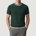 Man wearing a deep green knitted shirt with white and black striped cuffs, collar, and hem, hands in grey pants.