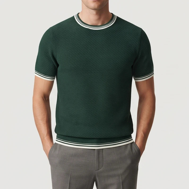 Man wearing a deep green knitted shirt with white and black striped cuffs, collar, and hem, hands in grey pants.