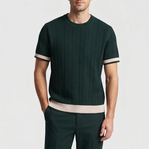 Man wearing a dark green cable knit short sleeve shirt with beige striped cuffs and hem, hands in pockets.